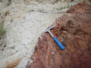 Detailbild: in der linken Hälfte und im oberen Teil graues Gestein, rechte Hälfte und unten rotes Gestein; an der Gesteinsgrenze liegt ein Geologenhammer.