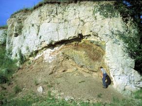 Felswand, die im oberen Bereich der Wand hellgrau ist; darunter ein Gestein mit grünen, gelben und roten Farben. Ein Mann steht als Größenvergleich rechts an der Wand: die Felswand ist etwa 4-mal so hoch wie er.