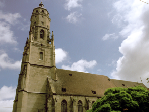 Die St. Georgskirche vor weiß-blauem Himmel, in der rechten unteren Bildhälfte ist eine riesige, grüne Baumkrone.