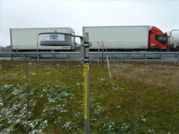 Das Bild zeigt im Vordergrund ein Plastikgehäuse, unter dem zwei Passivsammler angebracht sind. Das Plastikgehäuse dient dem Wetterschutz der Passivsammler und ist mit einem Winkel an einem Erdspieß befestigt, der im Boden verankert ist. Im Hintergrund sind Lkw‘s auf der Bundesautobahn A8 in der Nähe der Anschlussstelle Dasing bei Augsburg zu sehen.