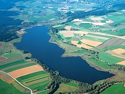 Das Luftbild zeigt den Silbersee in einer von Grünland und Ackerbau geprägten Landschaft des Oberpfälzer Waldes. Das Absperrbauwerk befindet sich am unteren Bildrand in der Mitte.