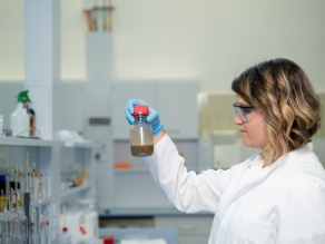 In einem Labor hält eine Frau im Labormantel eine Probenflasche, die zu einem Drittel mit etwas Braunem gefüllt ist, in die Höhe.