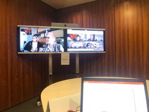 Zwei große Bildschirme in einer Ecke au denen eine Videokonferenz zu sehen ist.