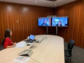 An einem großen Konferenztisch sitzt eine Frau am Laptop. In der Raumecke zwei große Bildschirme auf denen je eine Person zu sehen ist.