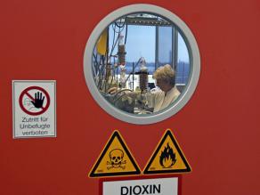 Aufnahme des Dioxin-Labors des LfU, sichtbar ist durch ein Bullauge eine Laborangestellte, der Schriftzug Dioxin sowie ein Totenkopf und eine Flamme als Gefahrensymbole