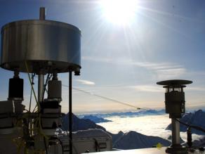 Aufnahme von zylindrischen Kartuschen, durch die Luft gesaugt wird, um Schadstoffe auf geeignetem Material anzureichern. Im Hintergrund die winterlichen Berge süd-östlich der Zugspitze unter blauem Himmel, während die Täler von einer Nebelschicht bedeckt sind.