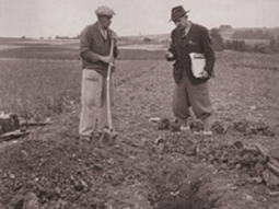 Schwarz-Weiß-Foto: Bodenkartierung der 1950er Jahre in Bayern - zwei Mitarbeiter nehmen ein Bodenprofil auf einem Acker auf und sprechen die Bodenart an.