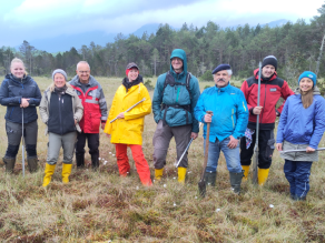 In einer offenen Hochmoorfläche stehen neun Männer und Frauen in Outdoor-Bekleidung und Gummistiefeln. In den Händen halten sie Schaufeln und anderes Werkzeug.