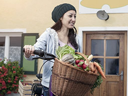Vor einem Haus steht eine Frau, die ihr Fahrrad am Lenker festhält. Daran ist ein Flechtkorb befestigt, der mit verschiedenen Gemüsesorten befüllt ist.