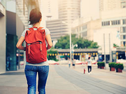 Eine Frau mit Rucksack, die von hinten zu sehen ist, geht eine Straße in einer Stadt entlang.