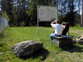 Zwei Personen sitzen auf einer Bank aus Stein und blicken auf eine Informationstafel.