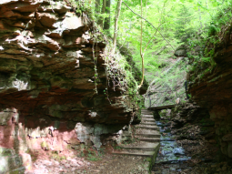 Wanderweg teilweise entlang des Bachs mit rotbraunen teils überhängenden Felswänden zu beiden Seiten.