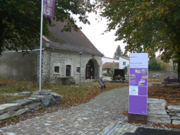  Haus steht im Hintergrund in mitten von Bäumen umgeben von Wiese und Wegen. Im Vordergrund ist ein menschengroßes Informationsschild zu sehen.