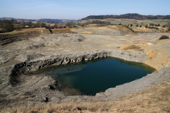 Tonabbau mit einer Wasserfläche im Vordergrund, aufgehaldetes Abraummaterial in der Grube und der Abbaukante im Hintergrund.