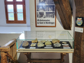 Ausschnitt eines Raumes, in dem auf einem Steinboden eine Tischvitrine mit Glashaube steht. In der Vitrine liegen 10 verschiedene Suevite und dazu eine kurze Erklärung über die Entstehung.