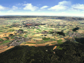Das Nördlinger Ries von oben als Luftbild, die Landschaft ist im Vordergrund bewaldet, dann folgen beige- und grün-farbige, meist rechteckige Landwirtschaftsflächen mit vereinzelten Orten unter weiß-blauem Himmel.