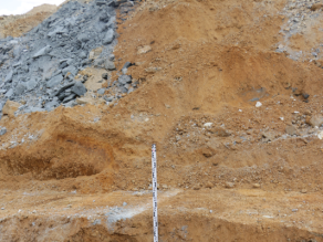 Geologische Schichten einer Bentonitgrube. An dem offenen Profil im Vordergrund liegt eine Messlatte an, und im unteren Teil ist eine graublaue Schicht Bentonit zu erkennen.