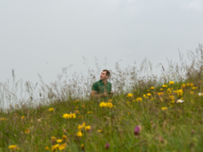 Ein Mann steuert mit einer Fernbedienung eine Drohne und verschwindet dabei in einer reich blühenden Bergwiese.