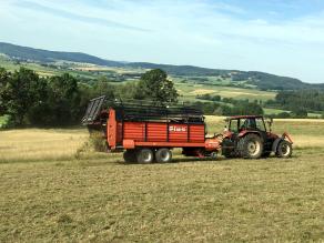 Im Vordergrund bringt ein Traktor mit Ladewagen Mähgut auf einer Grünlandfläche aus.