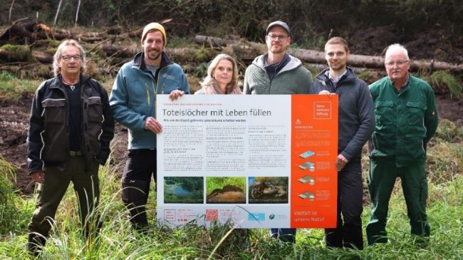 Eine Frau und fünf Männer stehen für einen symbolischen Spatenstich mitten im Wald auf Gilchinger Grund. Sie lächeln in die Kamera und halten ein großes Schild mit der Überschrift „Toteislöcher mit Leben füllen“ in der Hand.