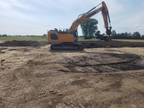 Ein orangener Bagger steht auf einer Fläche mit offenem braunem Erdboden und baggert einen Tümpel aus, im Hintergrund sind dunkelgrüne Gehölze vor einzelnen Wiesen zu sehen unter einem grau-blauem Himmel. 