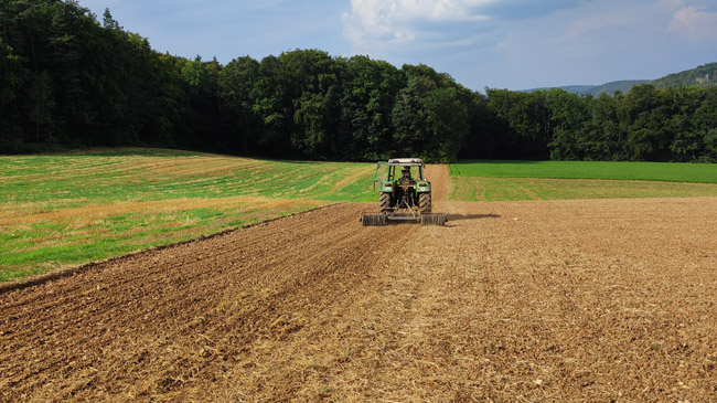In der Mitte des Bildes ist ein Traktor, der einen neu eingesäten Feldrain walzt. Links und rechts davon sind Ackerflächen nach der Ernte. Im Hintergrund ist Wald und darüber blauer Himmel mit weißen Wolken.