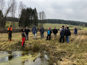 Die Naturpark-Rangerin erklärt einer Gruppe freiwilliger Helfer die Naturschutzmaßnahmen auf der Patenfläche. Sie stehen sich auf einer herbstlichen Wiese mit kleiner Feuchtfläche gegenüber und sind winterlich gekleidet. Im Hintergrund sind Bäume und dunkler Fichtenforst zu sehen.
