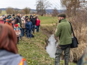 Ein Mann in güner Jacke steht mit dem Rücken zur Kamera neben einem Bach. Links im Bild steht eine Gruppe unterschiedlich alter Menschen und Blickt den Mann an. Rechts im Bild sind Stäucher zu sehen, im Hintergrund Wiese und einzelne Gehölze. 