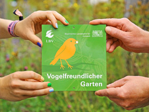 Vier Hände halten die Plakette 'Vogelfreundlicher Garten', die einen stilisierten gelben Vogel auf grünem Grund und die Logos des LBV sowie des LfU zeigt.
