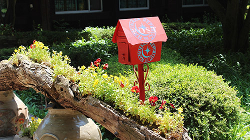Ein roter Briefkasten vor einem hohlen, mit Blumen bepflanzten Baumstamm.