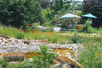 Naturgarten mit Mohnstauden, im Hintergrund Büsche, davor Gartenbänke, -tische und Sonnenschirme, im Vordergrund ein Teich, der mit Steinen abbegrenzt ist.