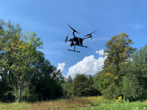 Fliegende schwarze Matrice 200-Drohne mit vier Propellern und montierter Wärmebildkamera in Bildmitte, umgeben von sommerlicher Parklandschaft bei blauem Himmel.