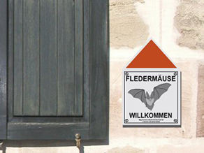 Plakette 'Fledermäuse willkommen' symbolisiert durch eine Fledermauszeichnung mit einem roten Dreieck darüber, an einer Hauswand