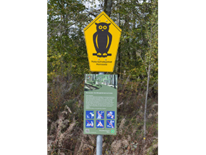 Schutzgebietsschild 'Eule' in schwarz auf gelben Grund, darunter eine Informationstafel mit Text zu dem Gebiet und Piktogrammen, die die wichtigsten Verbote und Gebote in plakativer Form darstellen.