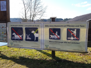 Eine direkt am Ufer eines Sees aufgestellte große Tafel mit plakativen Piktogrammen, die auf die konkrete Situation zu Verboten – wie Verbot von Wasserfahrzeugen aller Art, Hunde Baden verboten – und Geboten, wie Hunde anleinen, hinweisen.