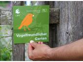 Die Auszeichnungsplakette der Aktion Vogelfreundlicher Garten wird von einer Person an einen Gartenzaun aus Holz gehalten