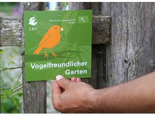 Die Auszeichnungsplakette der Aktion Vogelfreundlicher Garten wird von einer Person an einen Gartenzaun aus Holz gehalten