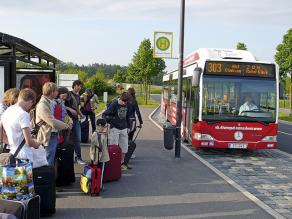Linienbus nähert sich einer Bushaltestelle, an der eine Gruppe von Menschen wartet
