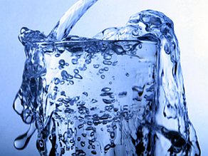 In ein volles Wasserglas vor blauem Hintergrund wird weiterhin schwungvoll Wasser gefüllt.