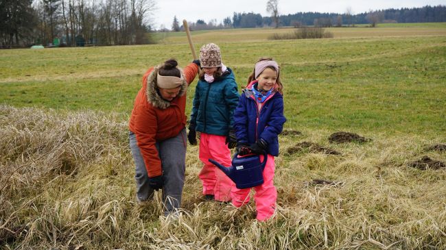Eine Frau pflanzt mit zwei Kindern einen Baum in einem Altgrasstreifen. Eines der Kinder trägt eine Gießkanne. Im Hintergrund Grünland mit vereinzelten Bäumen.