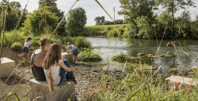 Zwei Personen sitzen auf einem Natursteinquader am abgeflachten Kiesufer eines Flusses. Im Hintergrund spielen zwei Kinder am Wasser.