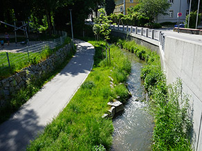 Blick von einer Brücke auf den Bach mit begrünten Ufern. Rechts daneben eine Ufermauer und eine höherliegende Straße. Auf der anderen Seite ein parallel verlaufender Geh- und Radweg.