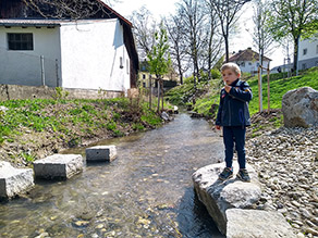 Ein Kind steht auf einem Felsen am Ufer eines Bachs und pustet Blumensamen in die Luft. Im Wasser befinden sich mehrere Trittsteine.
