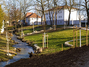 Ein leicht geschwungener Bach mit abgeflachten Ufern und Totholzelementen im städtischen Raum. Sitzreihen aus Natursteinquadern am Ufer und in der Böschung laden zum Verweilen ein. Ufer und Böschung wurden mit jungen Bäumen bepflanzt.