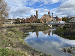 Der geschwungene Verlauf des neuen Seitenarms im Vordergrund. Ein Steg führt über das Gewässer. Im Hintergrund eine Kirche und die städtische Kulisse von Gunzenhausen.