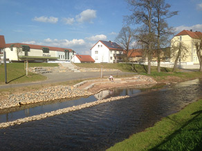 Auf einer Sitzsteinmauer am Ufer eines Flusses sitzt eine Person und schaut auf das Wasser. Im Hintergrund ein Geh- und Radweg und weitere Sitzmöglichkeiten in Form eines Atriums, welches in den Hochwasserschutzdeich integriert wurde. Auf einer Sitzsteinmauer am Ufer eines Flusses sitzt eine Person und schaut auf das Wasser. Im Hintergrund ein Geh- und Radweg und weitere Sitzmöglichkeiten in Form eines Atriums, welches in den Hochwasserschutzdeich integriert wurde.