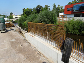 Eine Hochwasserschutzmauer am Ufer eines Flusses befindet sich im Bau. Stahlmatten stehen aus einer niedrigen Betonmauer heraus. Im Hintergrund lagerndes Baumaterial. Eine Hochwasserschutzmauer am Ufer eines Flusses befindet sich im Bau. Stahlmatten stehen aus einer niedrigen Betonmauer heraus. Im Hintergrund lagerndes Baumaterial.