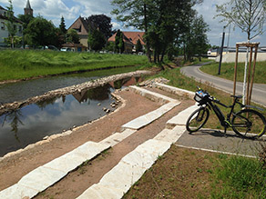Sitzstufen und eine Treppe führen die Böschung hinab zu einem Flussufer. Ein Fahrrad steht oberhalb davon neben einem Geh- und Radweg, der entlang des Flusses verläuft Sitzstufen und eine Treppe führen die Böschung hinab zu einem Flussufer. Ein Fahrrad steht oberhalb davon neben einem Geh- und Radweg, der entlang des Flusses verläuft