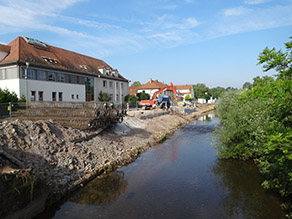 Am Ufer eines Flusses liegt Baustahl. Im Hintergrund wird die Verschalung der neuen Hochwasserschutzmauer im Bereich der Uferböschung erstellt. Ein Bagger und Lastkraftwagen stehen dahinter. Am Ufer eines Flusses liegt Baustahl. Im Hintergrund wird die Verschalung der neuen Hochwasserschutzmauer im Bereich der Uferböschung erstellt. Ein Bagger und Lastkraftwagen stehen dahinter.