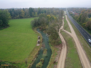 Luftaufnahme des Mühlbachs im Bereich des Hochwasserschutz Freilassing: Im Bach zwei kleine Inseln. Die Bachufer sind im Vordergrund überwiegend mit Gras, im Hintergrund mit Gehölzen bewachsen. Rechts parallel zum Bach verlauft ein Kiesweg und der neue Hochwasserschutzdeich mit Deichkronenweg. Hinter dem Deich eine viel befahrene Landstraße. Links vom Bach Grünland und Wald. Luftaufnahme des Mühlbachs im Bereich des Hochwasserschutz Freilassing: Im Bach zwei kleine Inseln. Die Bachufer sind im Vordergrund überwiegend mit Gras, im Hintergrund mit Gehölzen bewachsen. Rechts parallel zum Bach verlauft ein Kiesweg und der neue Hochwasserschutzdeich mit Deichkronenweg. Hinter dem Deich eine viel befahrene Landstraße. Links vom Bach Grünland und Wald.